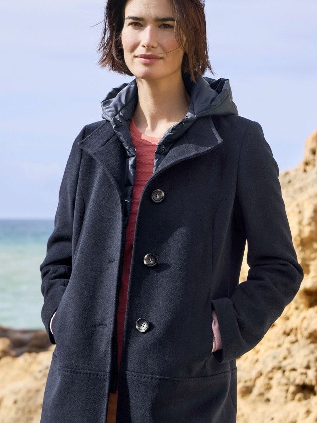 Ein Frau steht an einer Felsküste und trägt eine Wolljacke mit Steppeinsatz in Marine, darunter ein hellrotes Baumwollshirt.