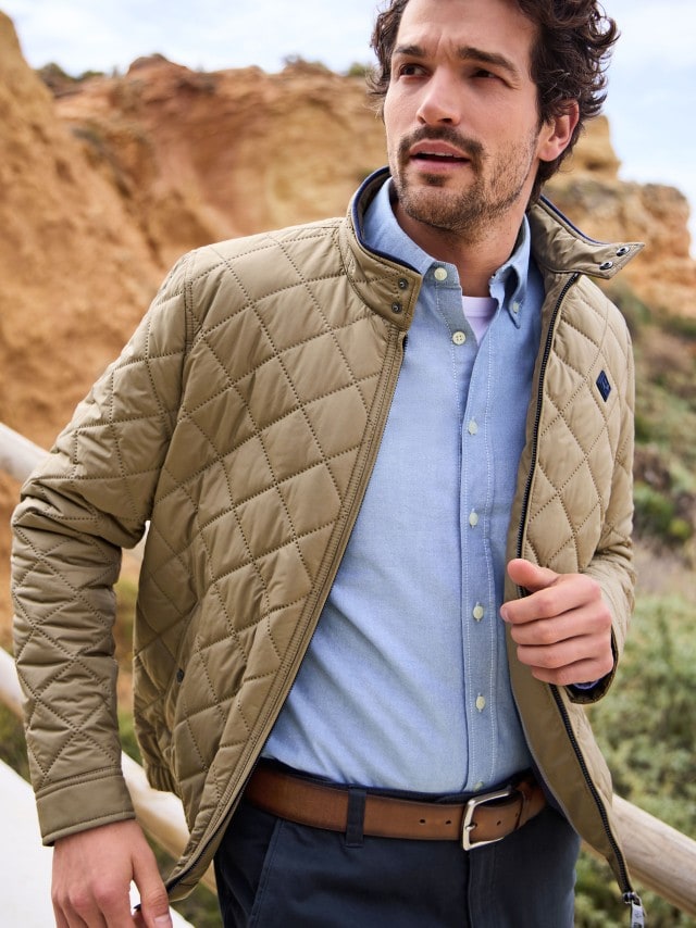 Ein Mann steht an einer Felsküste und trägt eine Steppjacke in Beige, darunter ein blaues Hemd und dazu eine Chino in Marine.