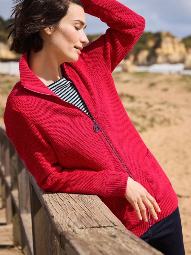 Frau in einer roten Strickjacke mit Reißverschluss über einem gestreiften Oberteil und Jeans lehnt an einem Holzzaun am Strand und schaut nach unten.