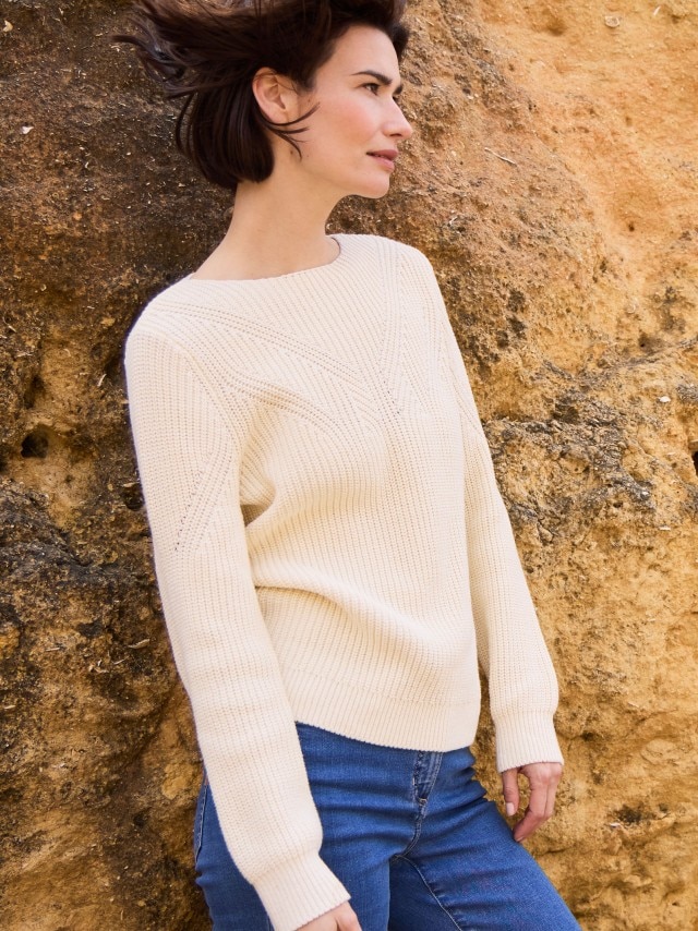 Eine Frau steht an einem Felsen und trägt einen Baumwoll-Pullover Patentstrick in Creme, dazu eine Jeans.
