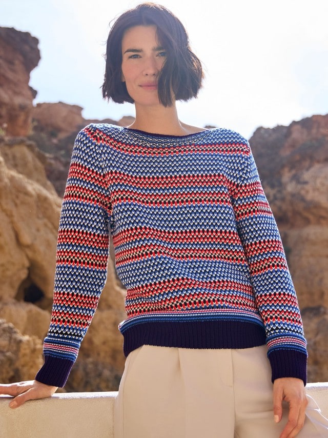 Eine Frau steht vor einem Felsen und trägt einen Baumwoll-Pullover Nordic Stripe, dazu eine Hose in Beige.