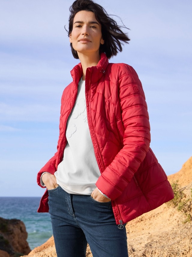Eine Frau steht an einer Küste und trägt eine rote Steppjacke Leichtgewicht, darunter ein Sweatshirt und dazu eine Jeans.
