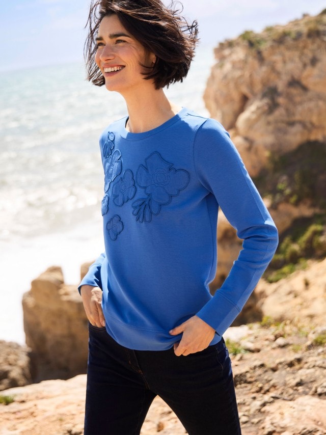 Eine Frau steht an einer Felsküste und trägt ein blaues Sweatshirt, dazu eine dunkele Jeans.