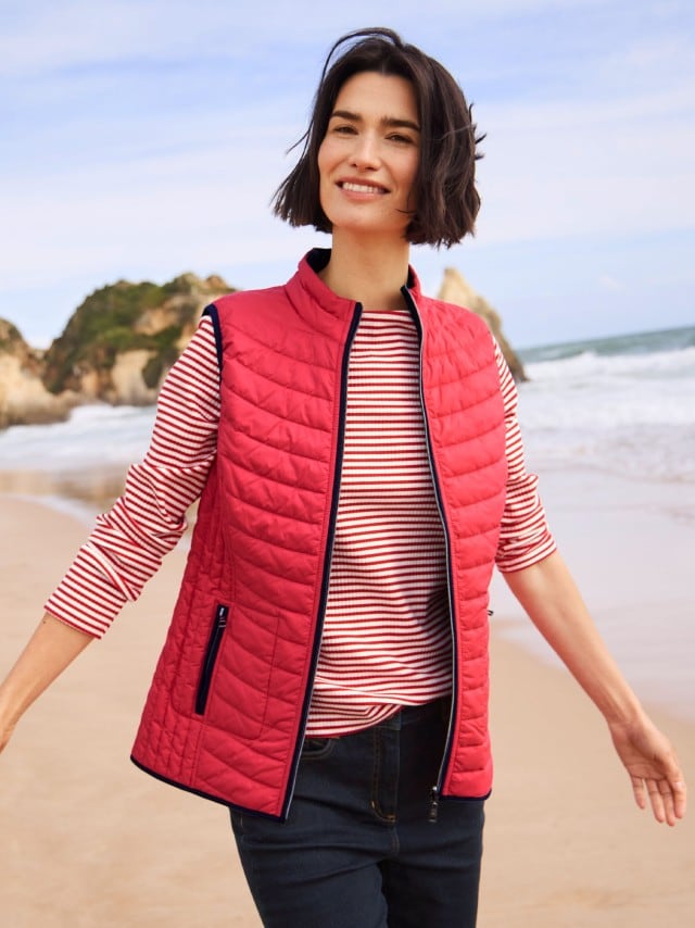 Eine Frau steht am Strand und trägt eine Wende-Steppweste in rot, darunter ein gestreiftes Langarm-Shirt und dazu eine Jeans. 
