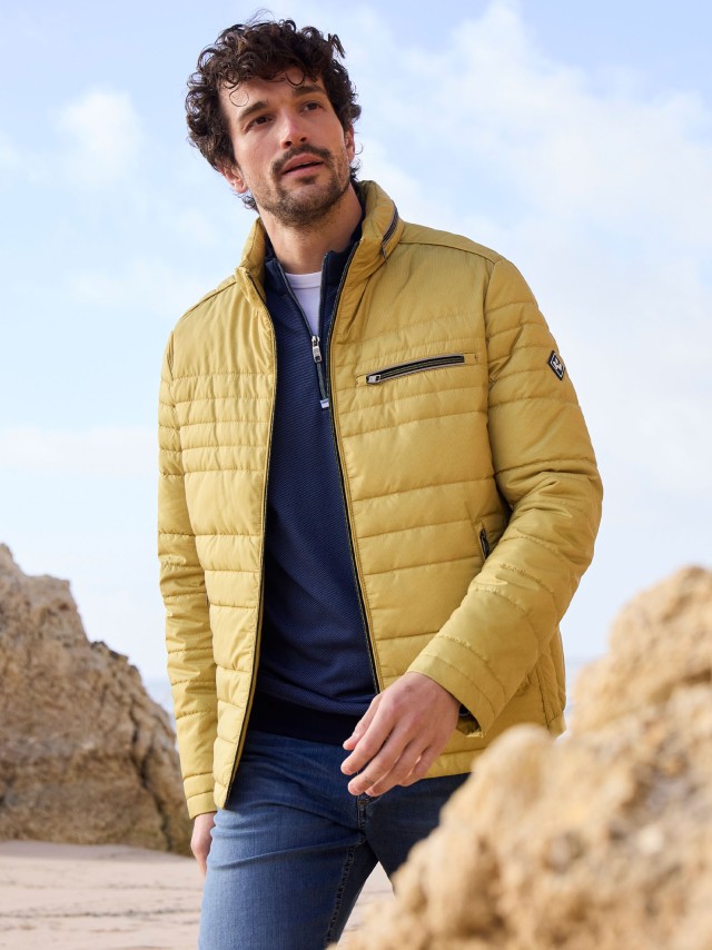 Ein Mann steht an einer Felsküste und trägt eine Microfaser Leichtjacke in Honiggelb, darunter einen Troyer und dazu eine Jeans.