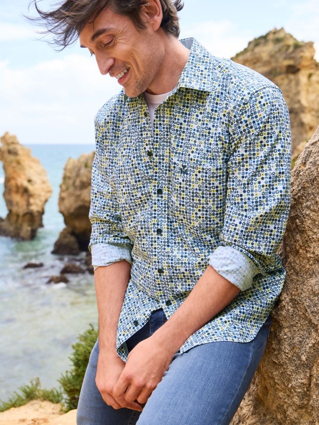 Ein Mann steht an einer Felsküste und trägt ein gemustertes Hemd, dazu eine helle Jeans.