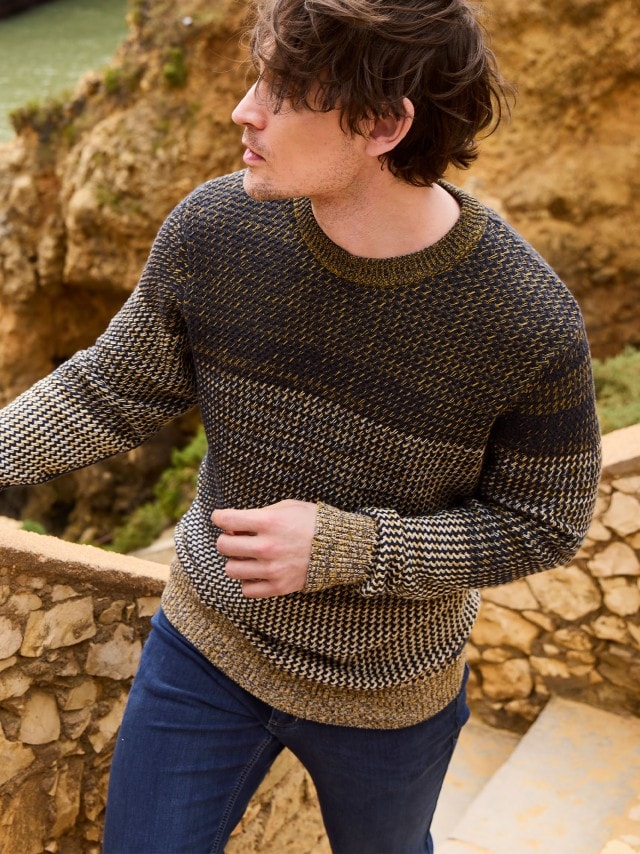 Ein Mann steht an einer Felsküste und trägt einen gestrickten Effekt-Pullover, dazu eine Jeans.
