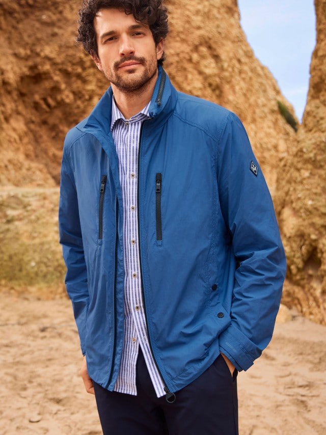 Ein Mann steht an einer Felsküste und trägt eine blaue Multifunktionsjacke, darunter ein gestreiftes Hemd und dazu eine Hose in Marine.