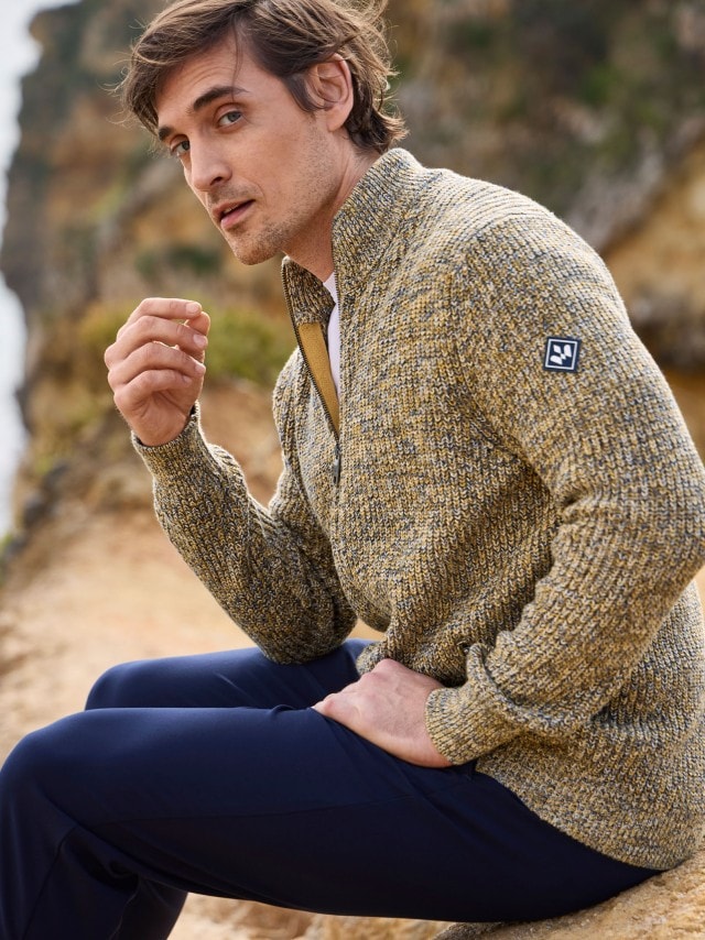 Ein Mann steht an einer Felsküste und trägt eine Ultrasonic Leichtsteppjacke in Beige, darunter ein blaues Hemd und dazu eine Chino in Marine.