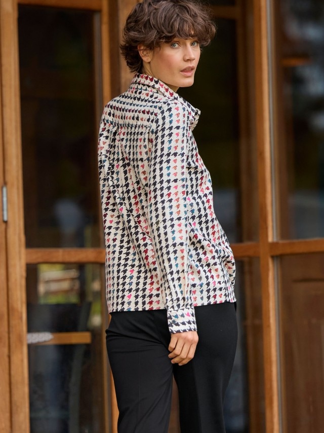 Eine Person trägt eine gemusterte Bluse im Hahnentritt-Stil mit bunten Akzenten. Kombiniert mit einer schwarzen Hose wirkt der Look modern und lebendig.