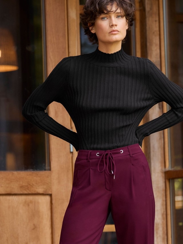 Eine Person trägt einen schwarzen, gerippten Pullover mit Stehkragen. Dazu kombiniert sind bordeauxrote Hosen mit Kordelzug, was einen modernen und eleganten Kontrast schafft.