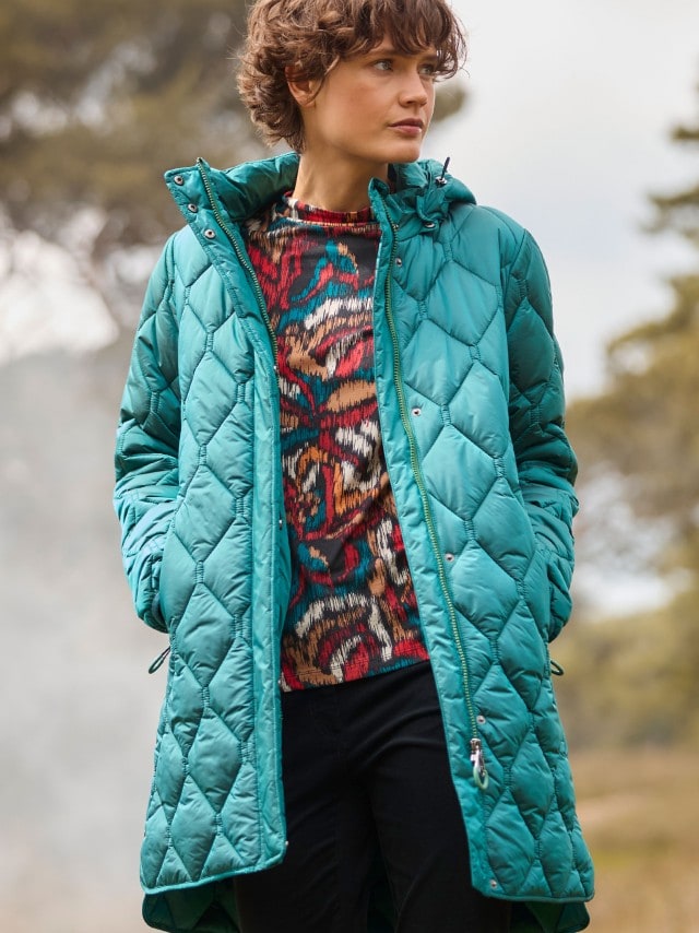 Eine Frau trägt einen langen, türkisfarbenen Steppmantel. Darunter ist ein bunt gemusterter Pullover kombiniert, was dem Look einen lebendigen und modernen Akzent verleiht.