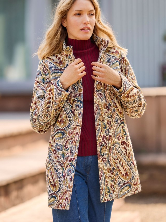 Eine Person trägt einen gemusterten Mantel im Brokat-Stil mit warmen Farbtönen. Darunter ist ein weinroter Rollkragenpullover kombiniert, was dem Look einen edlen und herbstlichen Charakter verleiht.