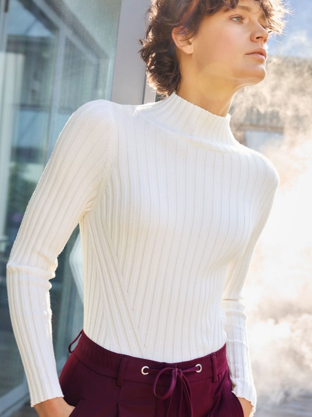 Eine Frau trägt einen weißen, gerippten Pullover mit Stehkragen. Dazu kombiniert sind bordeauxrote Hosen mit Kordelzug für einen klaren und modernen Look.