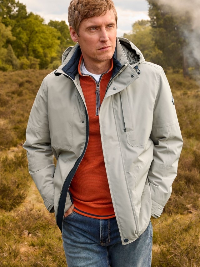Ein Mann steht im Freien und trägt eine hellgraue Outdoor-Jacke mit Kapuze über einem rostorangefarbenen Pullover sowie eine blaue Jeans.