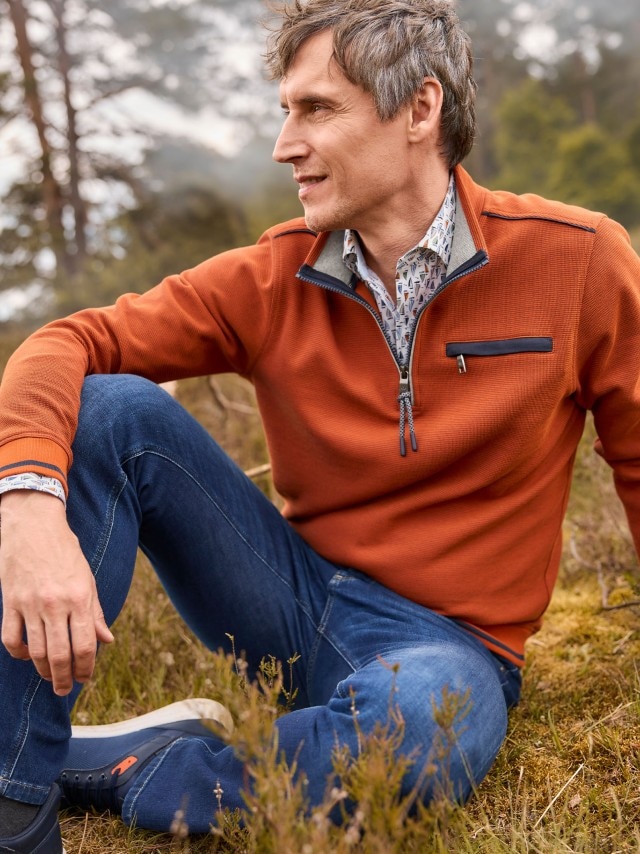 Ein Mann sitzt auf einer Wiese und trägt einen rostorangefarbenen Langarm-Pullover mit halbem Reißverschluss über einem gemusterten Hemd, dazu eine blaue Jeans und dunkelblaue Sneaker.