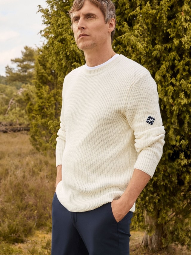 Ein Mann steht im Freien vor Bäumen und trägt einen weißen, grob gestrickten Pullover mit Abzeichen am Ärmel sowie eine dunkelblaue Hose.