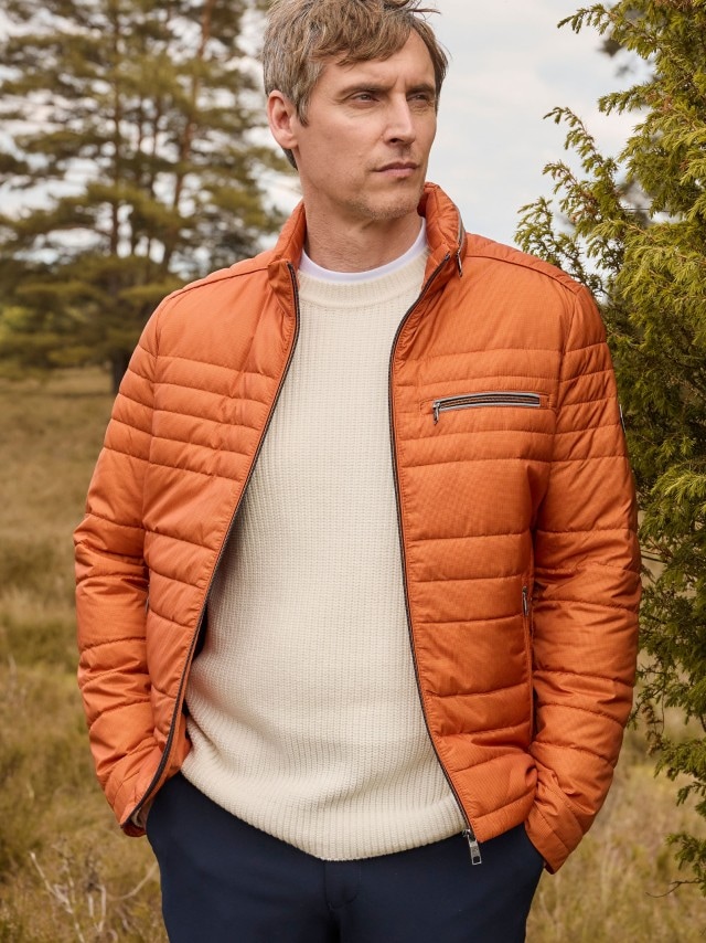 Ein Mann steht draußen vor Bäumen und trägt eine orange Steppjacke über einem hellen Strickpullover sowie eine dunkle Hose.