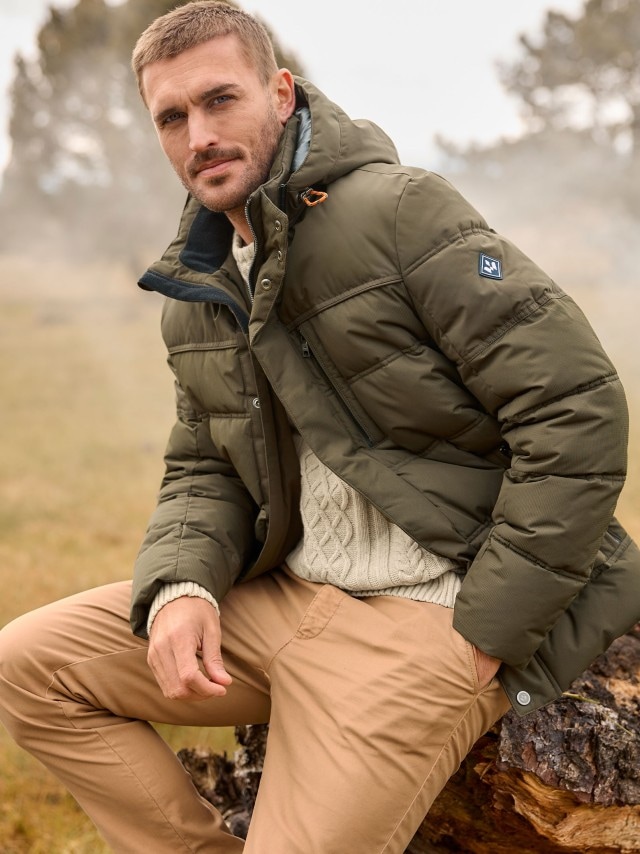 Eine Person sitzt im Freien auf einem Baumstamm und trägt eine olivgrüne Steppjacke mit Kapuze, einen beigen Strickpullover und eine hellbraune Hose.
