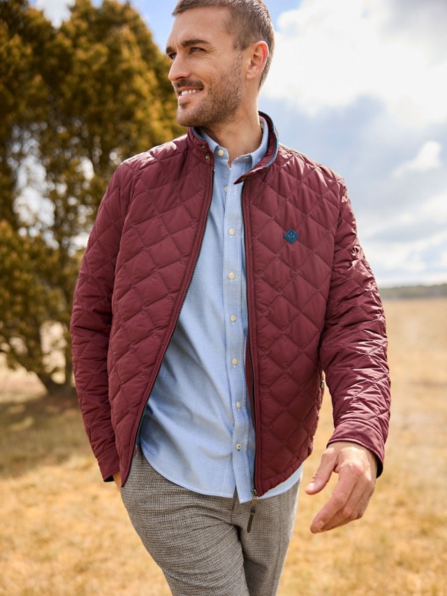 Eine Person geht im Freien und trägt eine weinrote, gesteppte Jacke über einem hellblauen Hemd und einer grau karierten Hose.