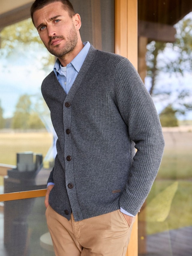 Eine Person steht mit den Händen in den Taschen und trägt eine graue Strickjacke über einem hellblauen Hemd sowie eine beige Hose.