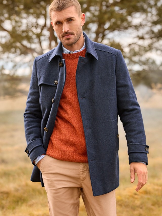 Eine Person steht im Freien und trägt einen dunkelblauen Mantel über einem orangefarbenen Pullover, kombiniert mit einem hellblauen Hemd und einer beigefarbenen Hose.