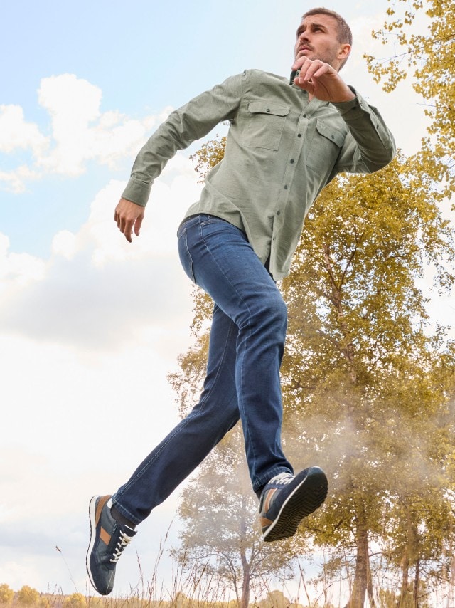 Eine Person springt im Freien und trägt ein hellgrünes Hemd mit Brusttaschen, blaue Jeans und dunkelblaue Sneaker mit braunen Akzenten.