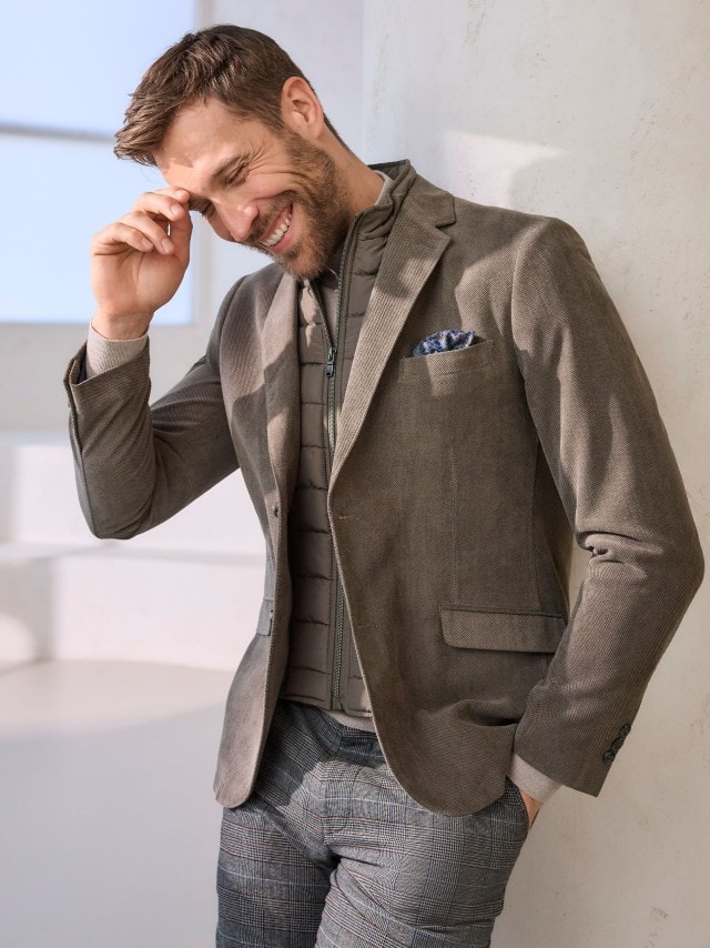 Mann in einem braunen Hemd und Sakko und grauer Anzughose lehnt an einer weißen Wand und schaut lächelnd in die Kamera.