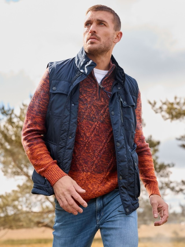 Eine Person steht im Freien und trägt eine dunkelblaue Steppweste über einem orange-roten Strickpullover mit Muster, kombiniert mit blauen Jeans.