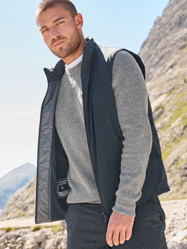 Ein Mann steht vor einem Berg und trägt eine Daunen Hybrid Weste in Marine, darunter einen grauen Pullover und dazu eine gestreifte Flanellhose.