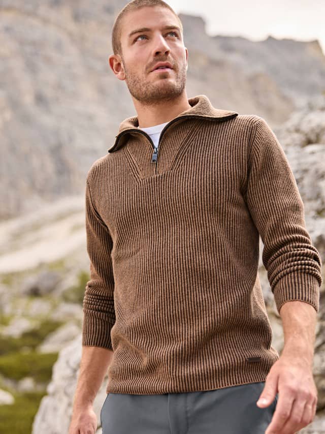 Ein Mann steht in einer felsigen Berglandschaft und trägt einen braun gestreiften Strickpullover mit halbem Reißverschluss über einem weißen T-Shirt sowie eine graue Hose.