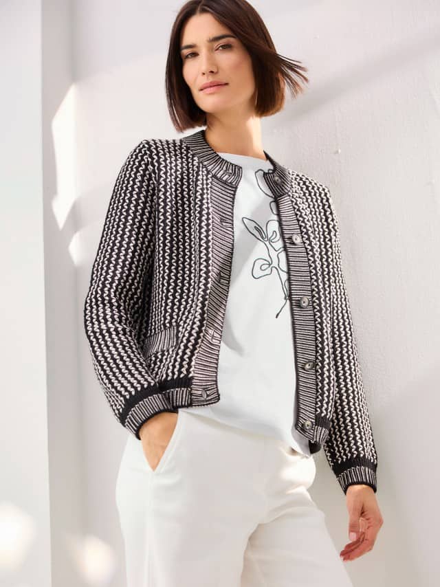 Eine Frau steht vor einer hellen Wand und trägt die Strickjacke Chic & Go in Schwarz/Creme, darunter ein Baumwollshirt mit Kordelstickerei und dazu eine helle Marlenehose mit Struktur.
