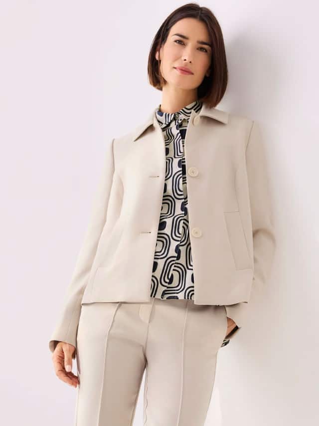 Eine Frau steht vor einer hellen Wand und trägt einen cremefarbenen Kofferblazer in A-Linie, dazu die passende Hose und dazu eine Baumwollbluse mit langer Manschette mit grafischem Muster. 