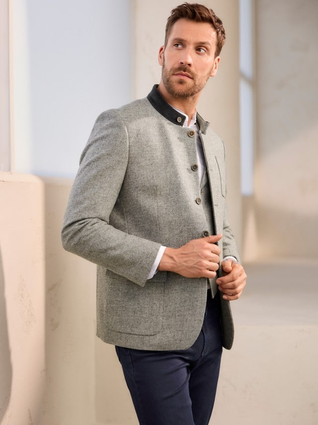 Ein Man trägt einen Lodenjanker, darunter ein weißes Hemd und dazu eine Hose in Marine. 