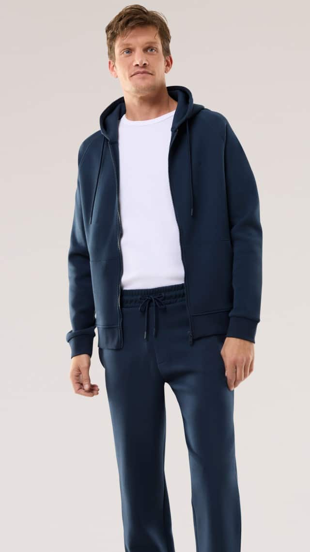 Ein Mann trägt einen dunkelblauen Freizeitanzug mit Hoodie und passender Hose, kombiniert mit einem schlichten weißen T‑Shirt. Der Look wirkt sportlich, modern und bequem.