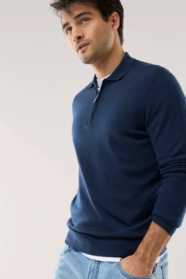 Ein Mann trägt einen dunkelblauen Pullover mit Kragen und halbem Reißverschluss sowie eine hellblaue Jeans. Der Look wirkt lässig und zugleich gepflegt.