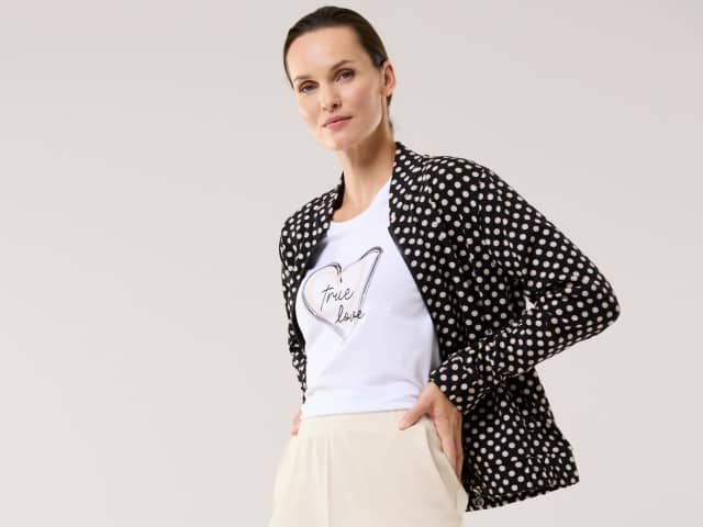 Eine Frau trägt eine schwarz‑weiß gepunktete Jacke über einem weißen T‑Shirt mit Herzmotiv und heller, weiter Hose. Der Look wirkt modern, lässig und leicht elegant.