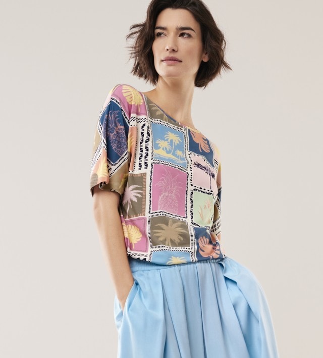 Eine Frau steht in einem hellen Raum, trägt ein bunt gemustertes Shirt und einen weiten hellblauen Rock. 