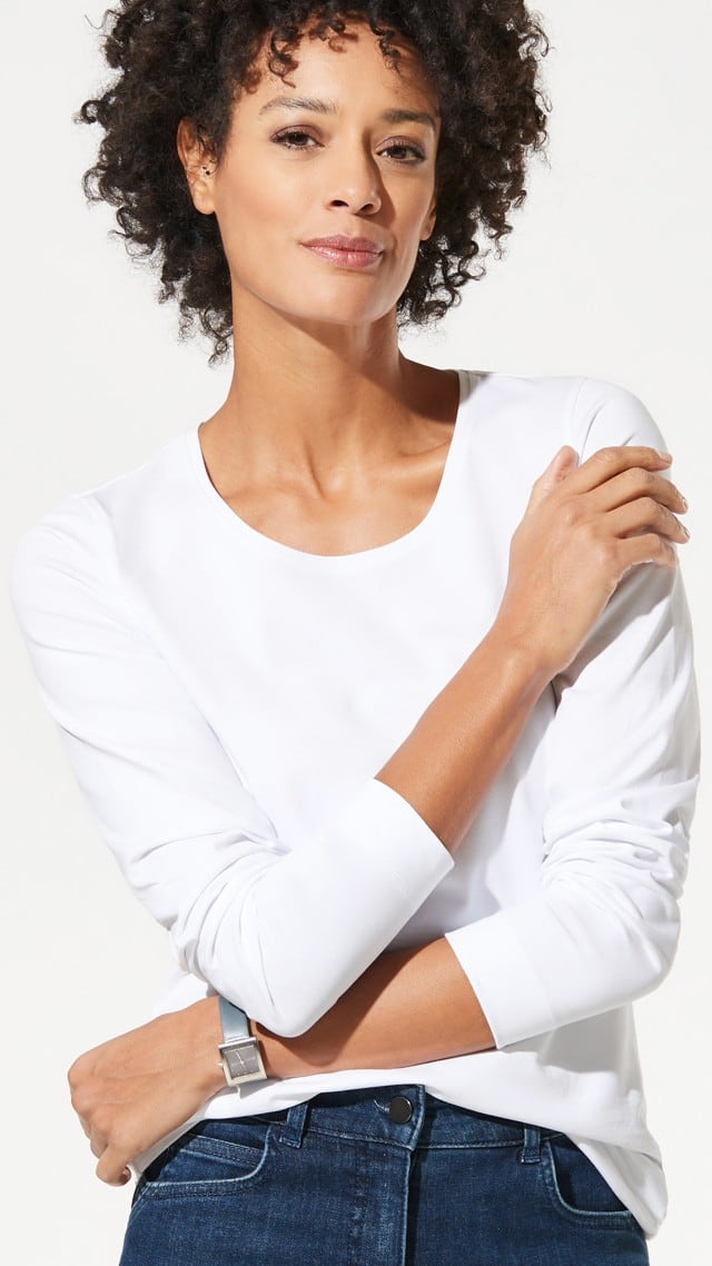 Eine Frau trägt ein schlichtes, langärmliges weißes Shirt und eine dunkelblaue Jeans. Der Look wirkt minimalistisch, modern und lässig.
