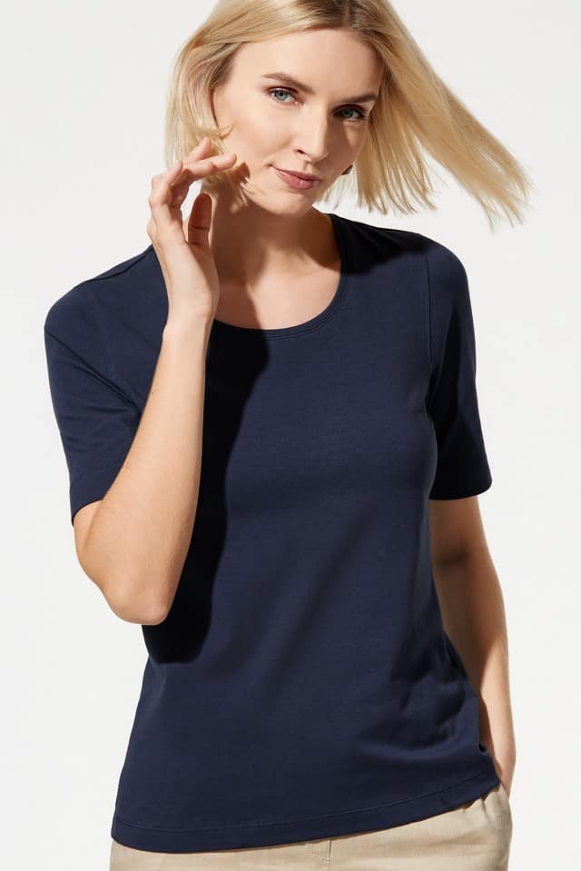 Eine Frau trägt ein dunkelblaues, kurzärmliges T-Shirt und eine hellbeige Hose. Das Outfit wirkt schlicht, modern und alltagstauglich.