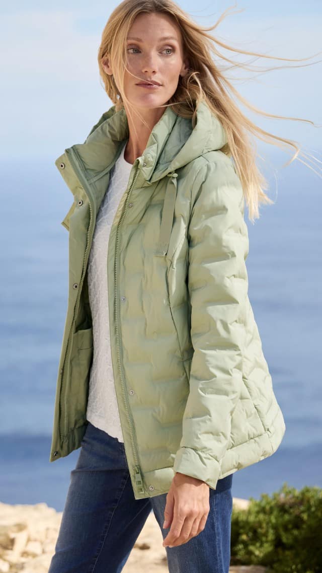 Eine Frau steht im Freien und trägt eine mintgrüne Steppjacke zu einem weißen Oberteil und Jeans.