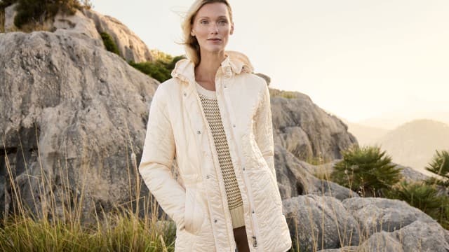 Eine Frau steht vor einer felsigen Landschaft und trägt eine helle Jacke über einem gestrickten Oberteil.