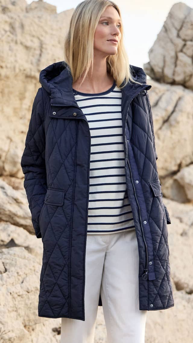 Eine Frau steht vor Felsen und trägt einen dunkel gestepp­ten Mantel über einem gestreiften Oberteil.