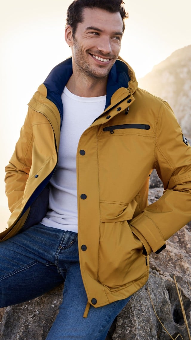 Ein Mann sitzt in einer Felslandschaft und trägt eine gelbe Jacke über einem weißen T‑Shirt und Jeans.