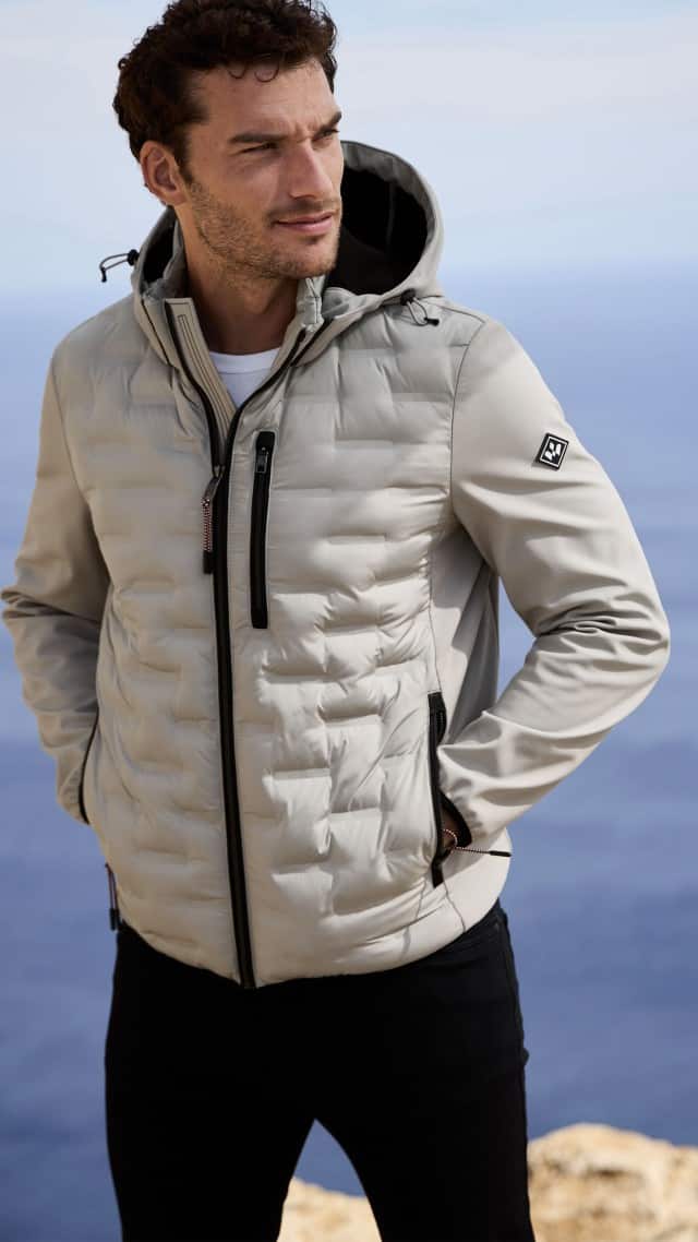 Ein Mann steht vor einem Meerblick und trägt eine hellgraue Kapuzenjacke mit schwarzen Reißverschlüssen.