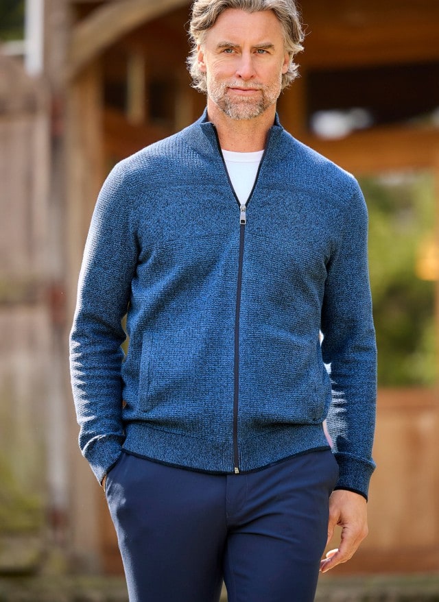 Ein Mann in einer blau-melierten Strickjacke und blauer Hose läuft locker vor eine Gebäude entlang und schaut zur Seite. Ein Mann in einer blau-melierten Strickjacke und blauer Hose läuft locker vor eine Gebäude entlang und schaut zur Seite.