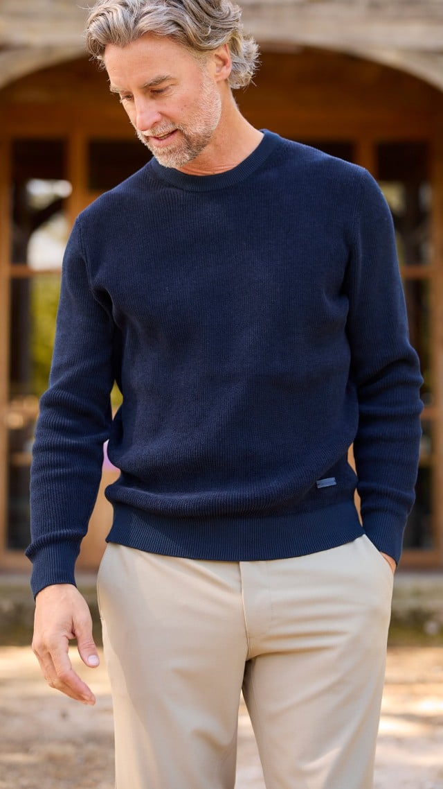 Ein Mann steht vor einem Gebäude, trägt einen dunkelbauen Ripppullover und eine helle Hose und schaut nach unten. Ein Mann steht vor einem Gebäude, trägt einen dunkelbauen Ripppullover und eine helle Hose und schaut nach unten.