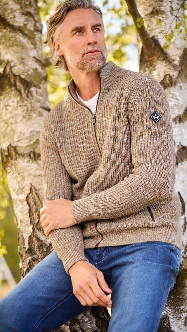 Ein Mann lehnt an einem Baum, trägt eine braun-melierte Strickjacke über einem weißen Shirt und Jeans und schaut zur Seite. Ein Mann lehnt an einem Baum, trägt eine braun-melierte Strickjacke über einem weißen Shirt und Jeans und schaut zur Seite.