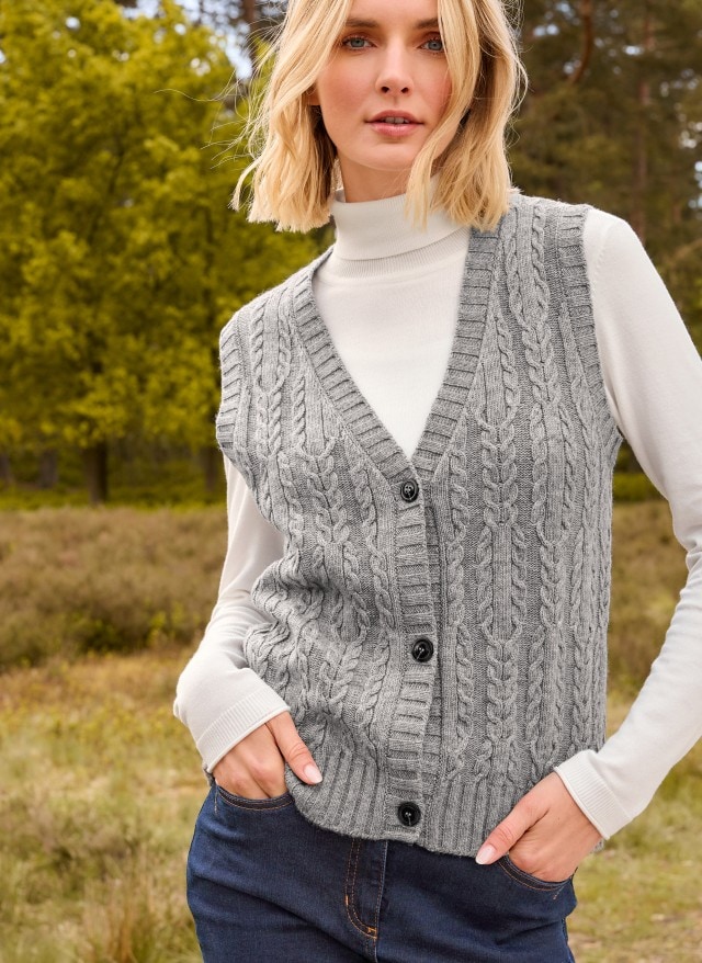 Eine Frau trägt einen weißen Rolli, darüber eine Strickweste in grau und dazu eine Jeans.