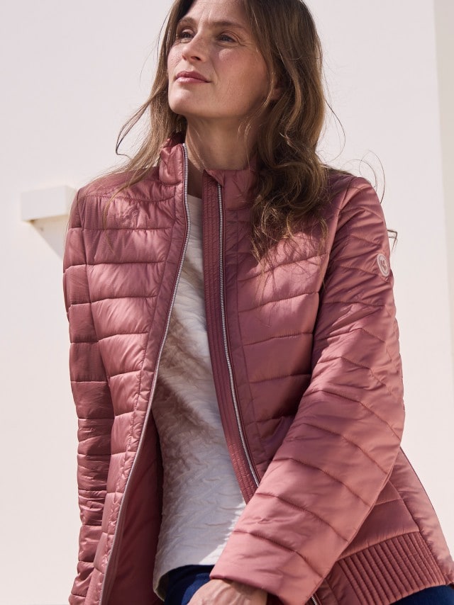 Eine Frau trägt die Leichtsteppjacke Komfort in der Farbe Rosenholz, darunter ein helles Shirt. 